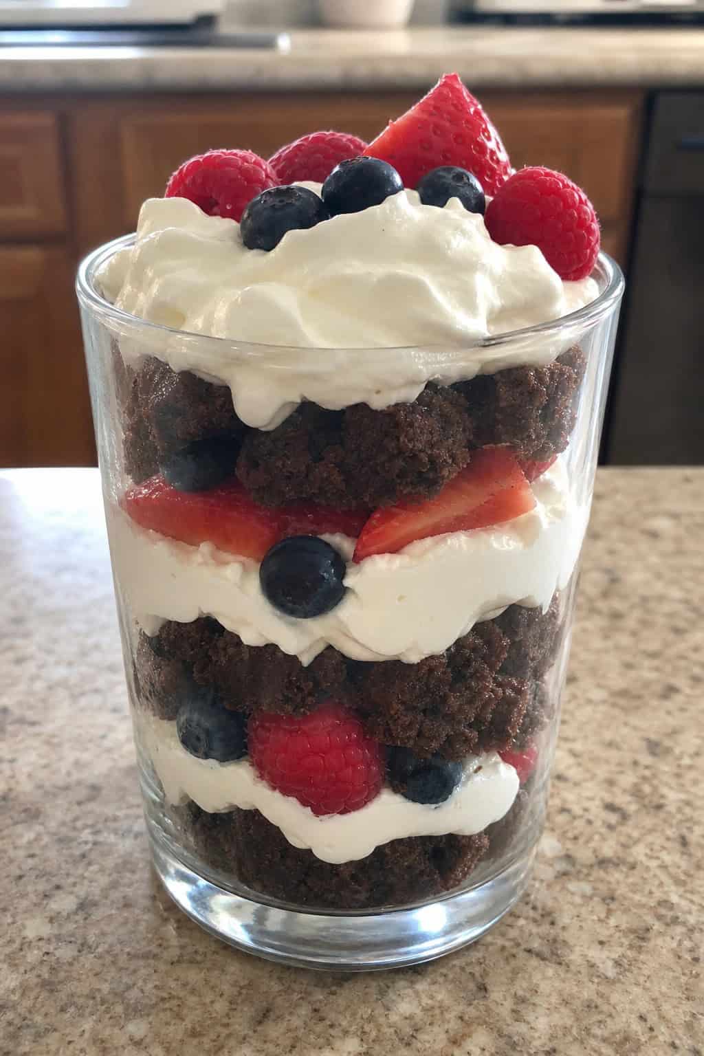 A clear glass parfait layered with brownie pieces, whipped cream, and mixed fresh berries on a wooden table.