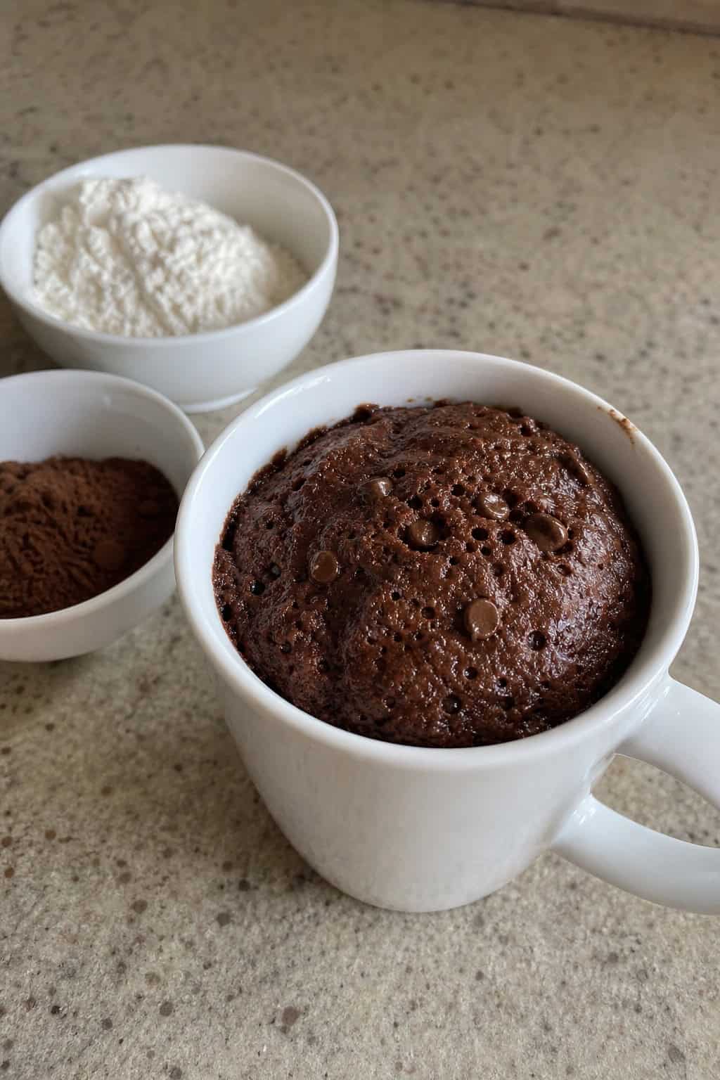 A single chocolate brownie mug cake in a white mug on a wooden table with baking ingredients around it.