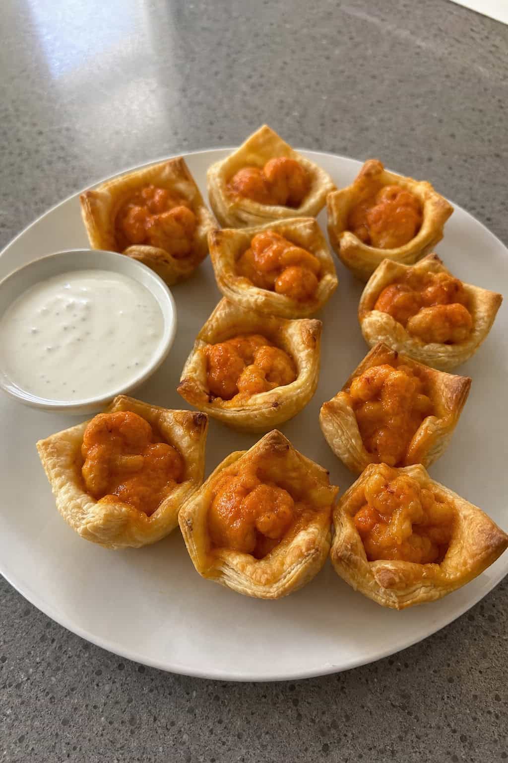 Buffalo cauliflower puff pastry wontons arranged on a wooden board with a bowl of dipping sauce and celery sticks.