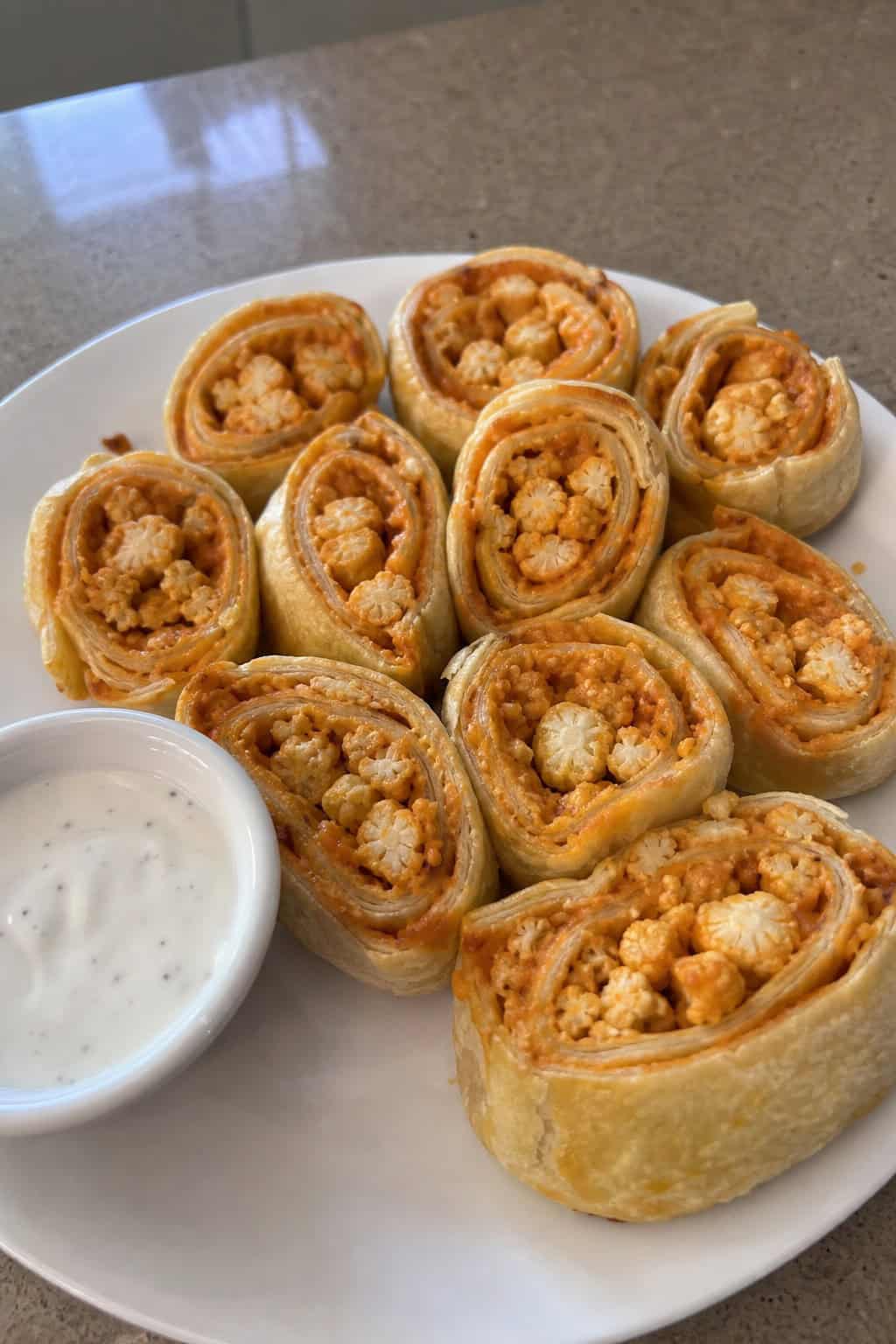 Close-up of buffalo cauliflower vegan pinwheels on a wooden board showing orange-sauced crispy cauliflower florets and shredded romaine inside spinach-tomato wrap tortillas, with a small dipping sauce bowl and celery sticks.