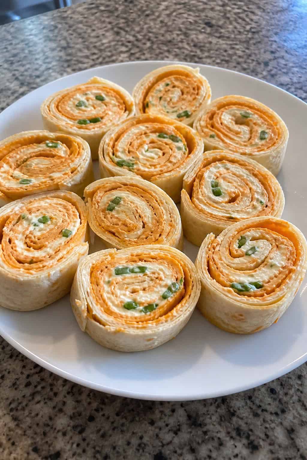 Buffalo chicken cream cheese pinwheels arranged on a wooden board showing orange-tinted filling with visible green onions, alongside small bowls of dipping sauces.