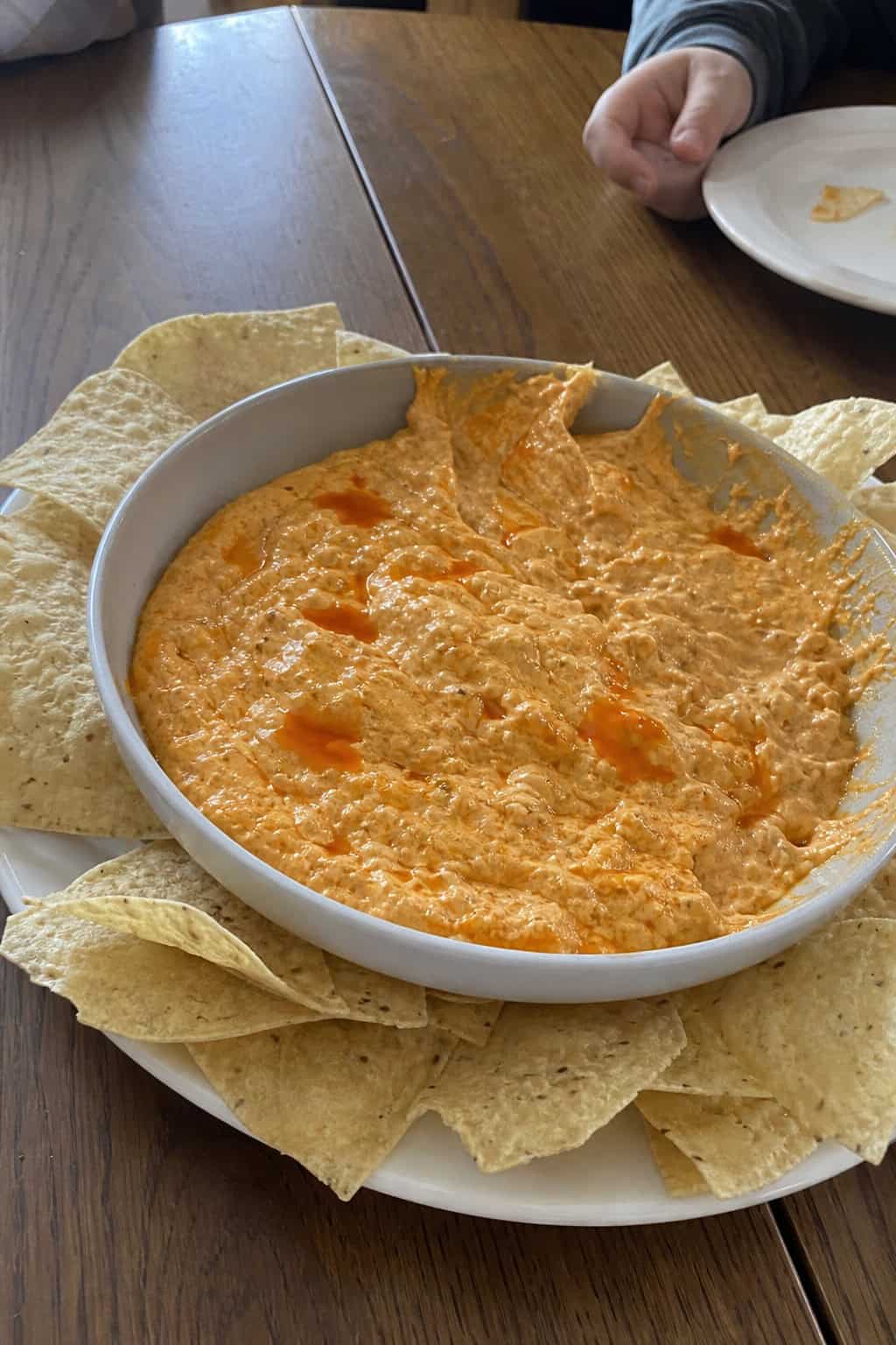 A bowl of buffalo chicken dip with tortilla chips and celery sticks on a wooden table, with a blurred family game night setup in the background.
