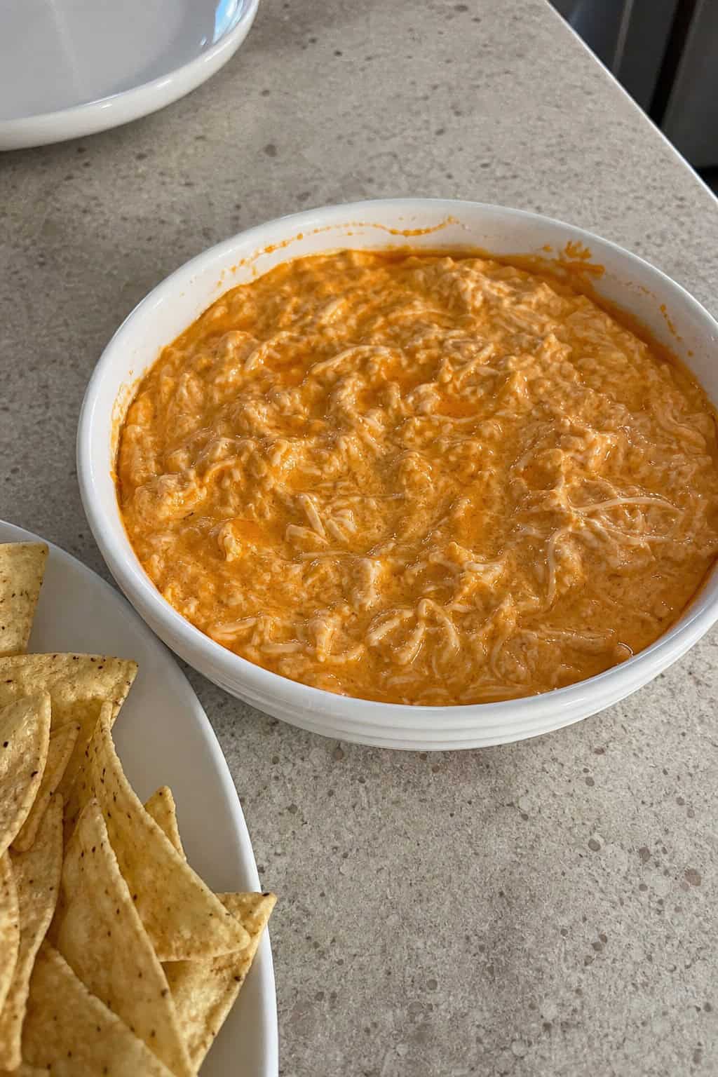A bowl of creamy buffalo chicken dip with shredded chicken, surrounded by celery sticks, carrot sticks, and tortilla chips on a wooden board.