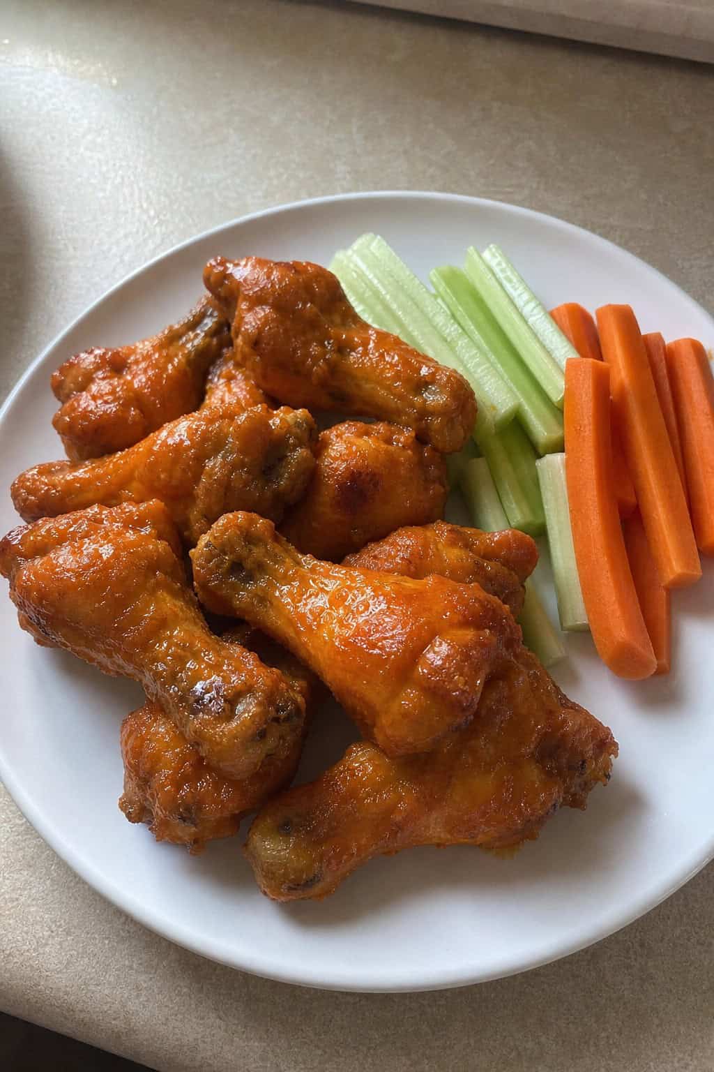 Caramelized buffalo-sauced chicken drumsticks arranged on a wooden serving board beside raw celery sticks and thin carrot batons.