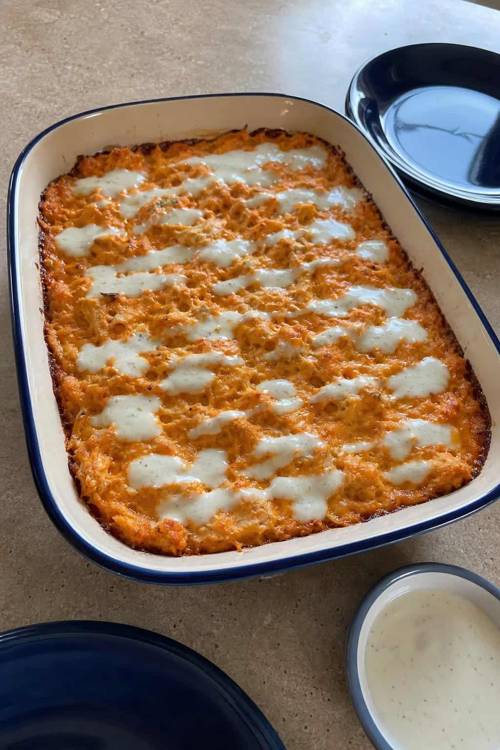 A baked buffalo chicken hashbrown casserole topped with cheese and green onions on a wooden table with bowls of sauce and celery sticks nearby.