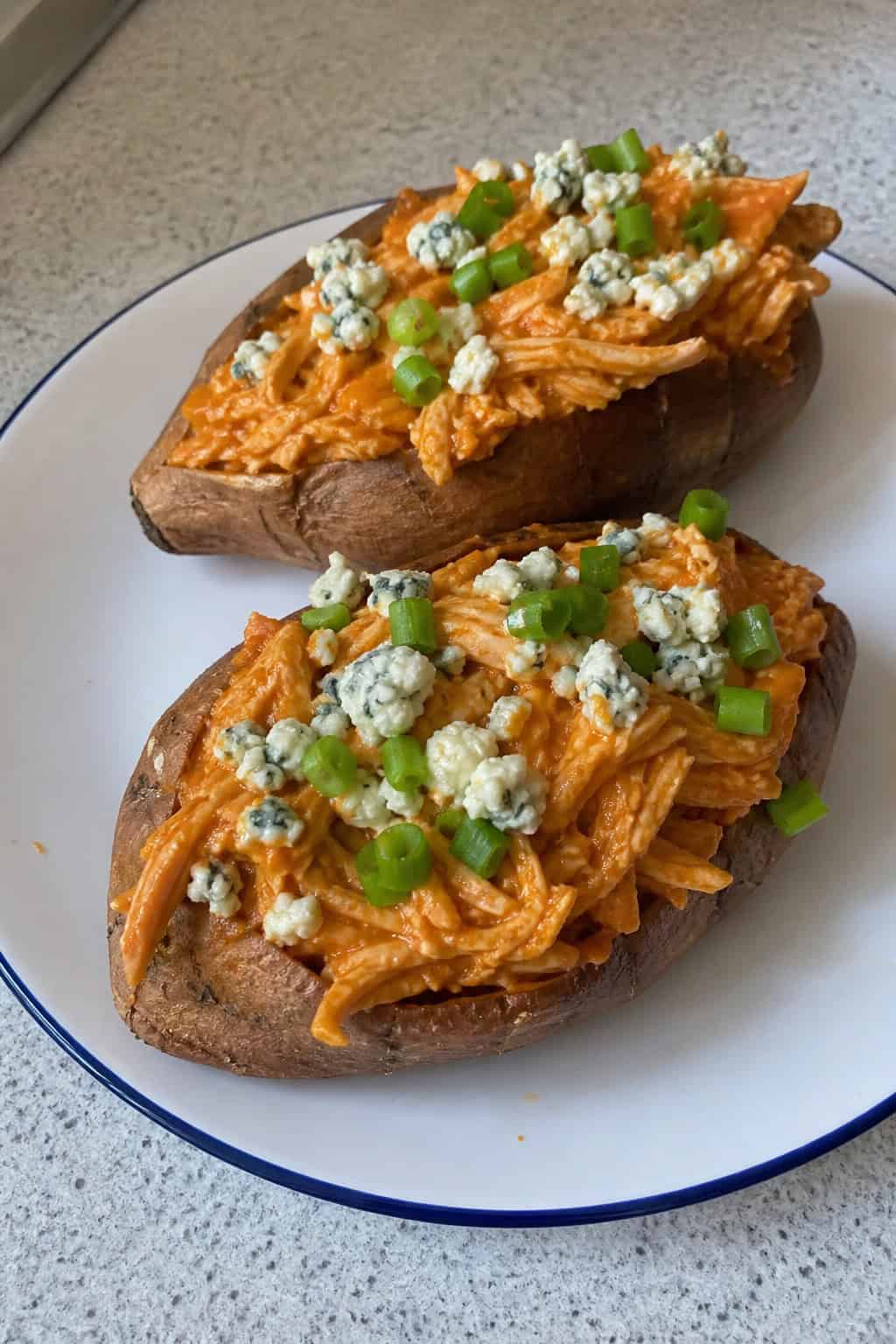 Two baked sweet potatoes split open and stuffed with shredded buffalo chicken, topped with crumbled blue cheese and sliced green onions, on a wooden table with celery sticks and a small bowl of sauce nearby.