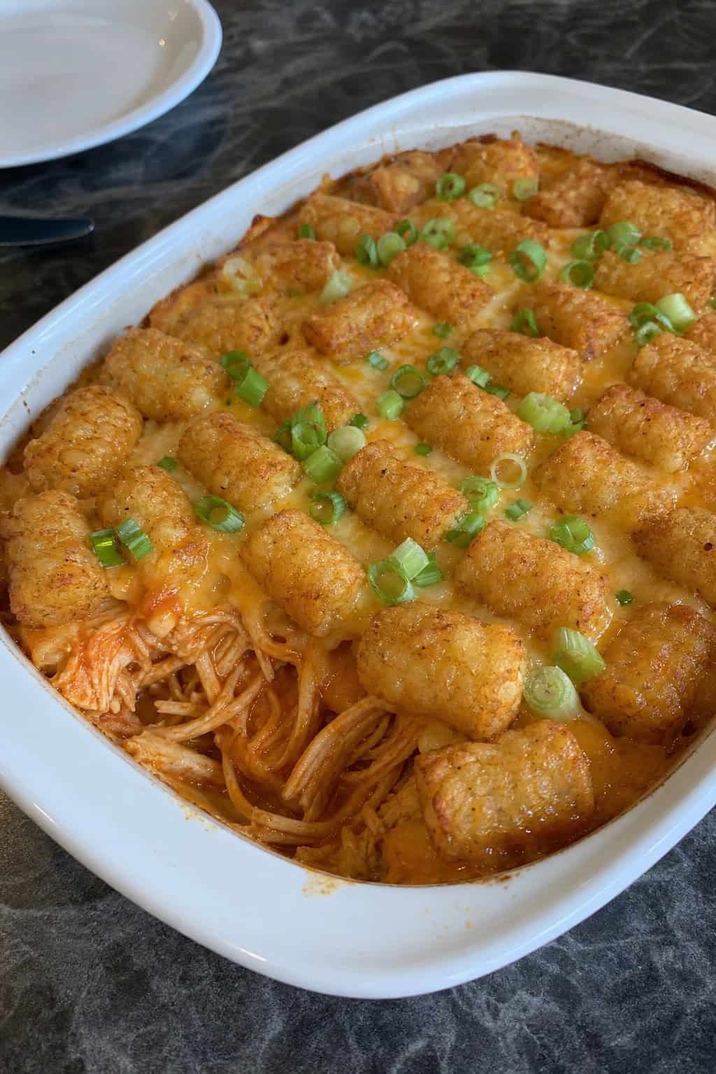 A baked Buffalo chicken tater tot casserole in a cream ceramic dish with golden tots on top, saucy shredded orange-tinged chicken visible at the sides, and sliced green onions scattered across the surface.