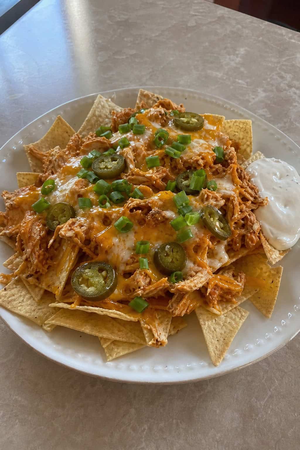 A plate of buffalo ranch canned chicken nachos topped with melted cheese, jalapeños, green onions, and cilantro, served with ranch dressing and lime wedges on a wooden table.