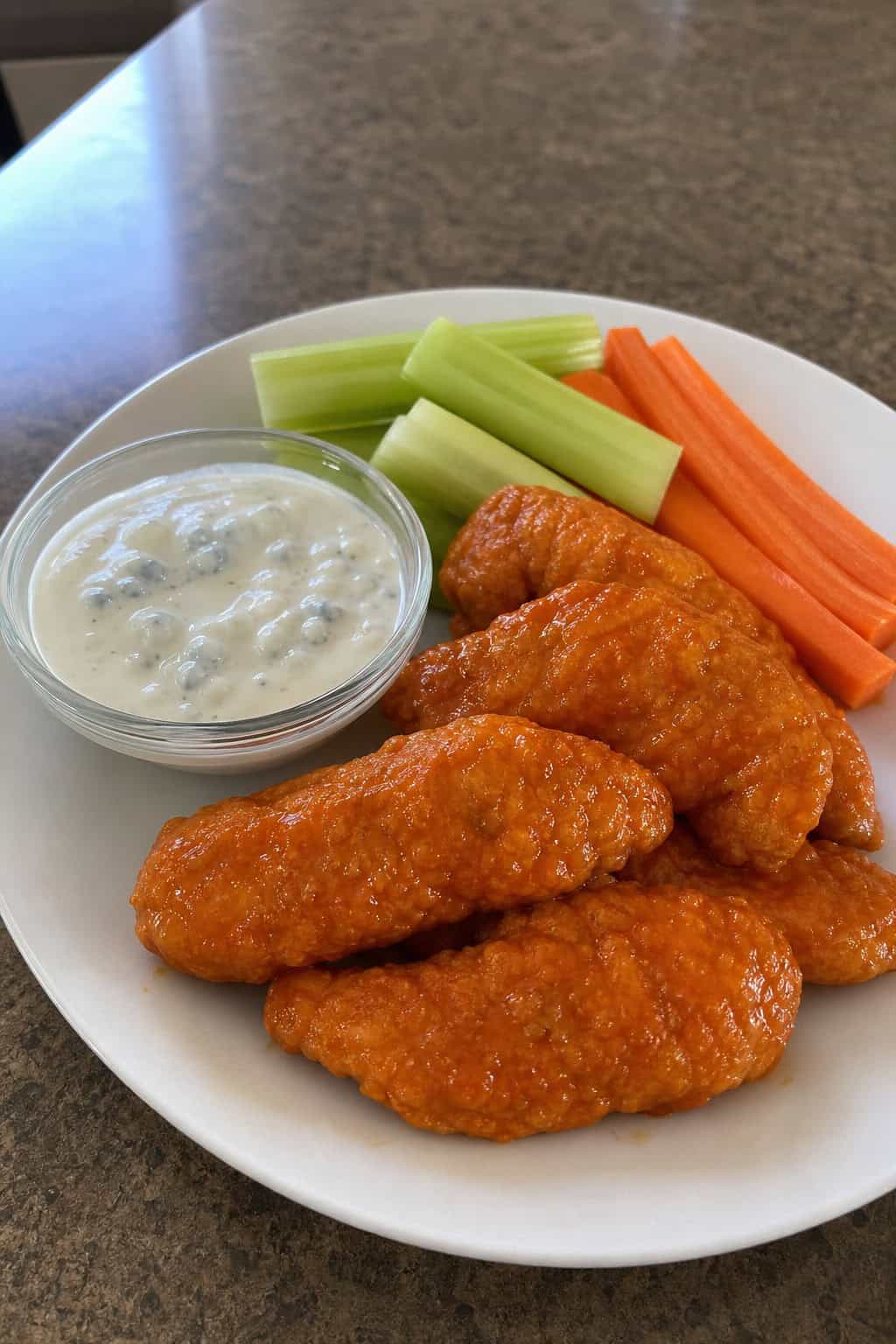 Plate of buffalo ranch chicken tenders with a bowl of blue cheese dip and fresh celery and carrot sticks on a wooden table.