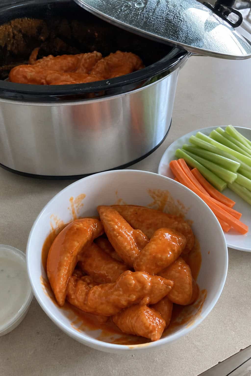 A bowl of saucy buffalo-coated chicken tenderloins served with celery sticks and carrot sticks, with a slow cooker visible in the background.