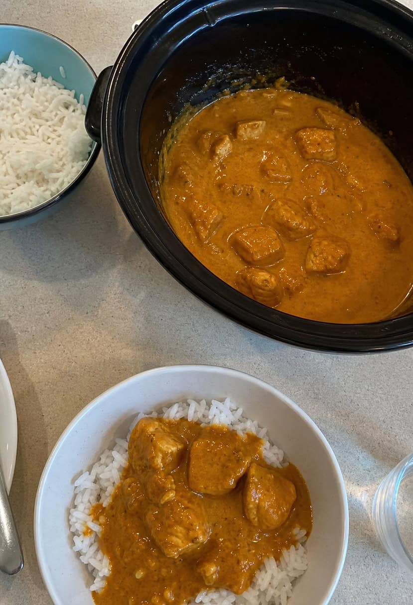 A crockpot filled with simmering butter chicken, surrounded by ingredients and spices on a kitchen counter