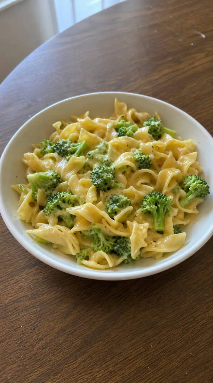 A bowl of shell pasta in a pale cheddar sauce with small soft broccoli pieces folded throughout, served in a white bowl on a wooden table