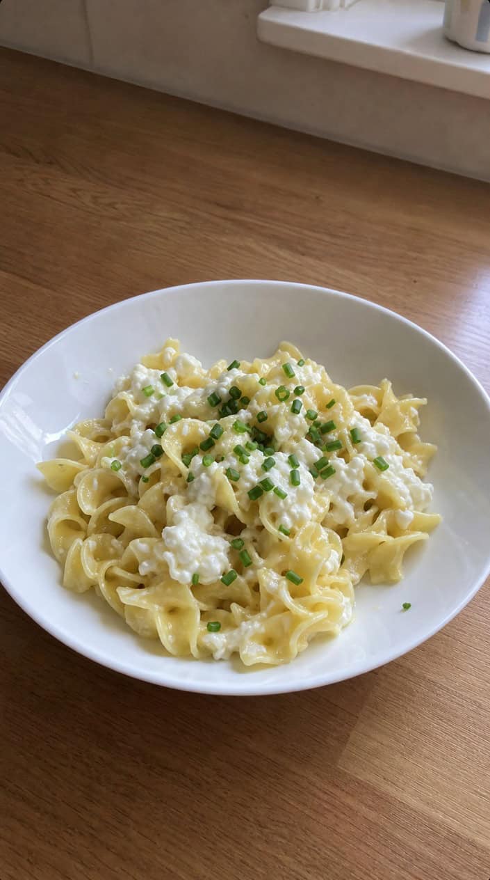 A bowl of spaghetti noodles folded with small-curd cottage cheese and flecks of green chives, sitting on a light wooden table