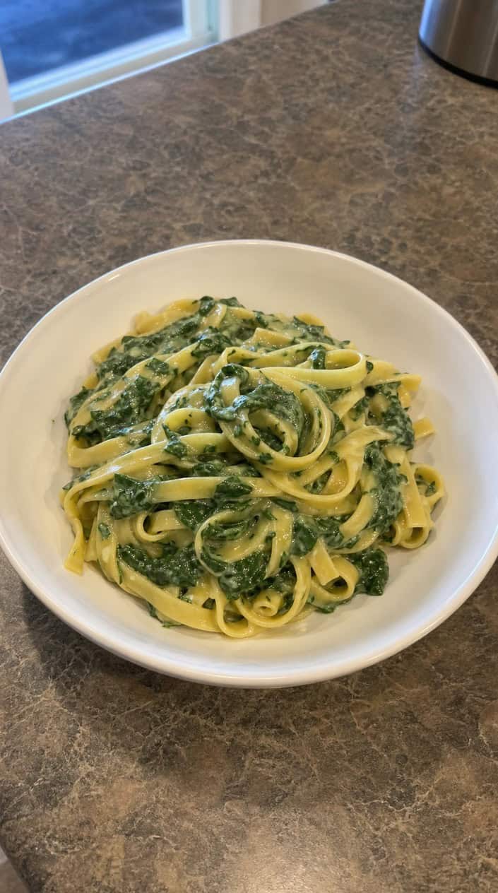 A white bowl of egg noodles with a pale green creamed spinach swirl across one side, a silver fork resting beside it on a wooden table