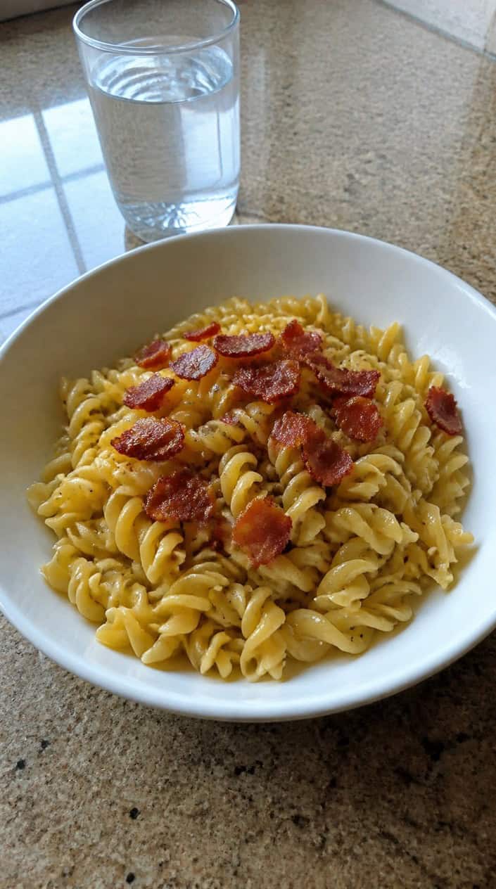 A bowl of wide egg noodles glistening with butter and scattered with dark crispy bacon crumbles, served on a plain wooden table