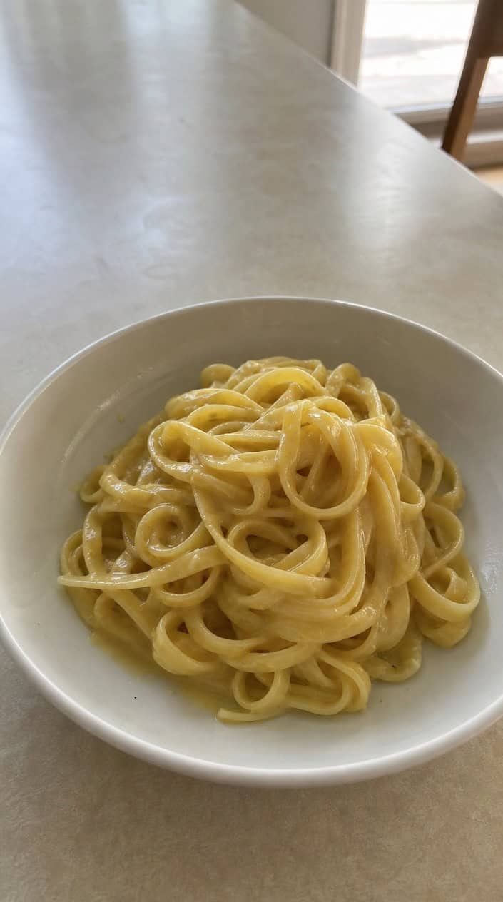 A ceramic bowl of pale yellow buttered noodles with a smooth creamy coating, sitting on a worn wooden table with a small grater in the background