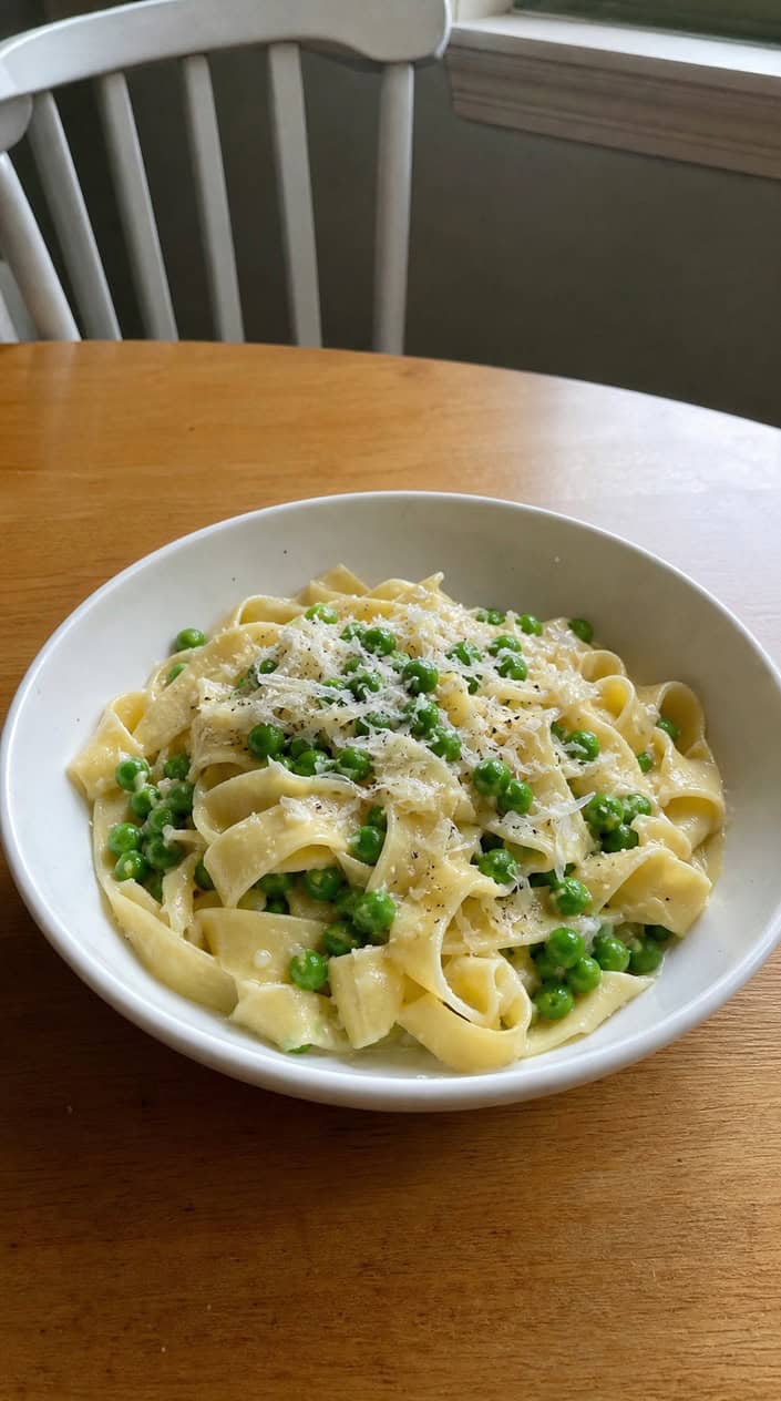 A bowl of buttered egg noodles dotted with bright green peas and a snow of grated Parmesan, photographed on a light wooden surface