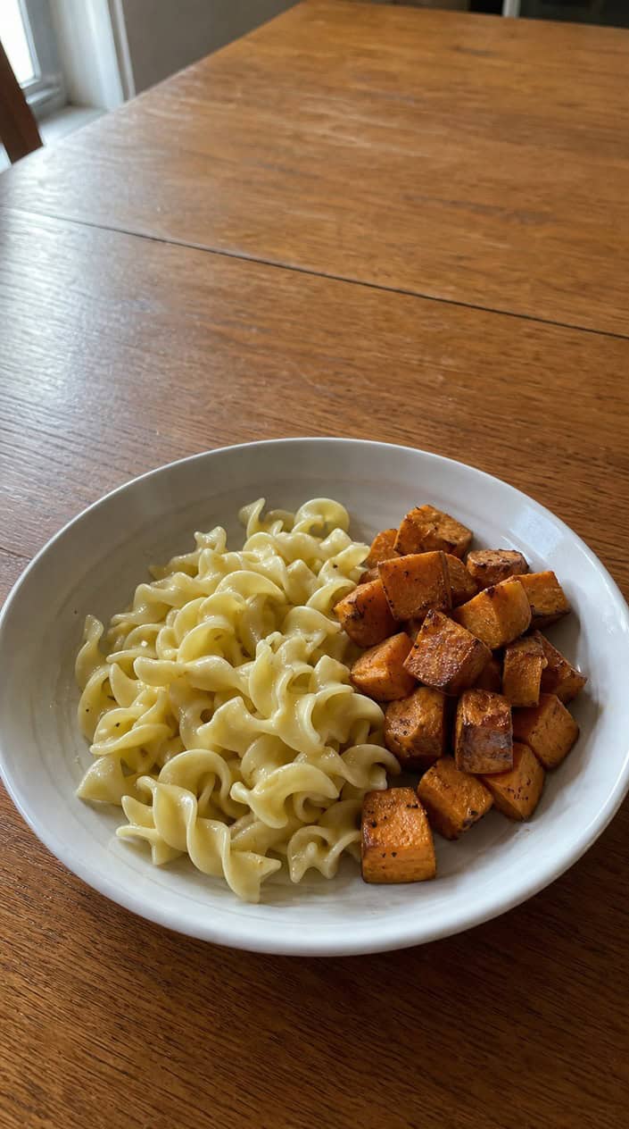 A bowl of elbow pasta in glossy butter sauce studded with deep orange roasted sweet potato cubes, with a fork and folded napkin resting on the wooden table beside it