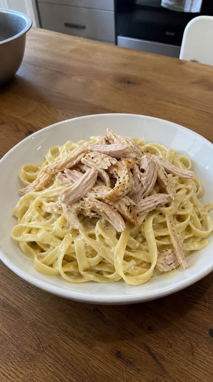 A wide bowl of golden egg noodles topped with loosely pulled rotisserie chicken strands, set on a warm wooden table
