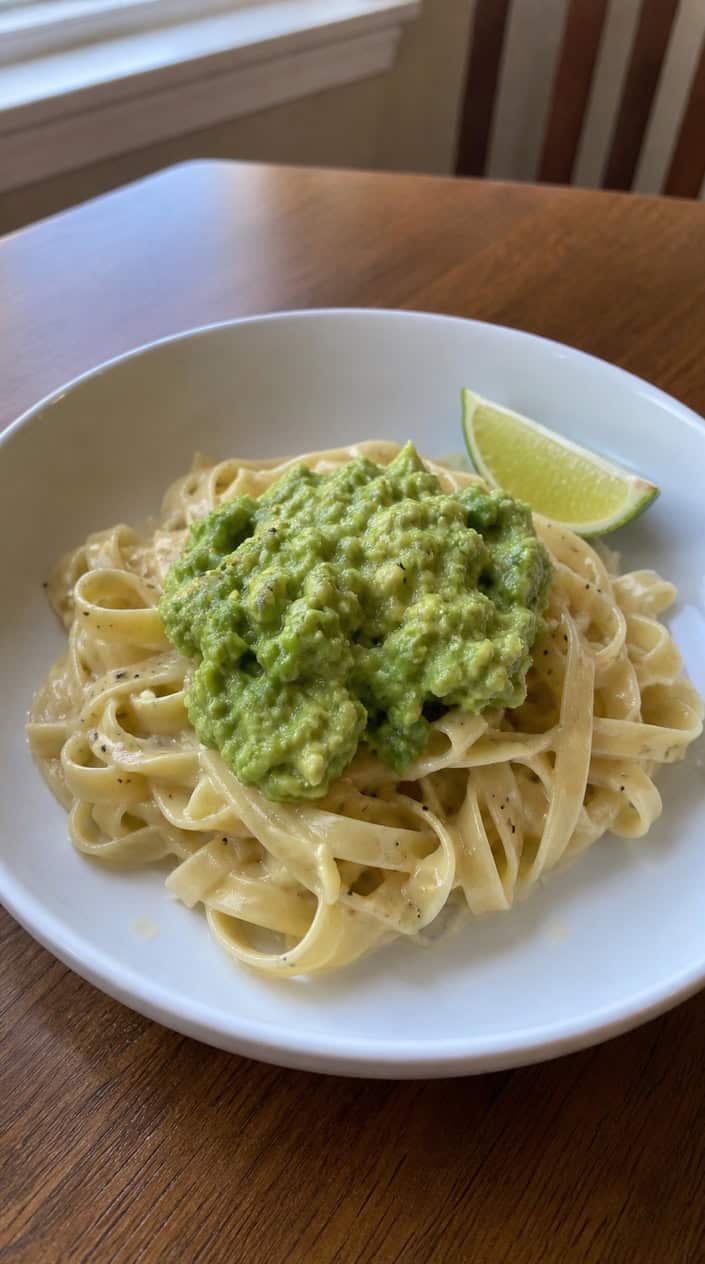 A bowl of small shell pasta coated in green-tinted avocado butter with a fresh lime wedge resting on the rim of a white bowl