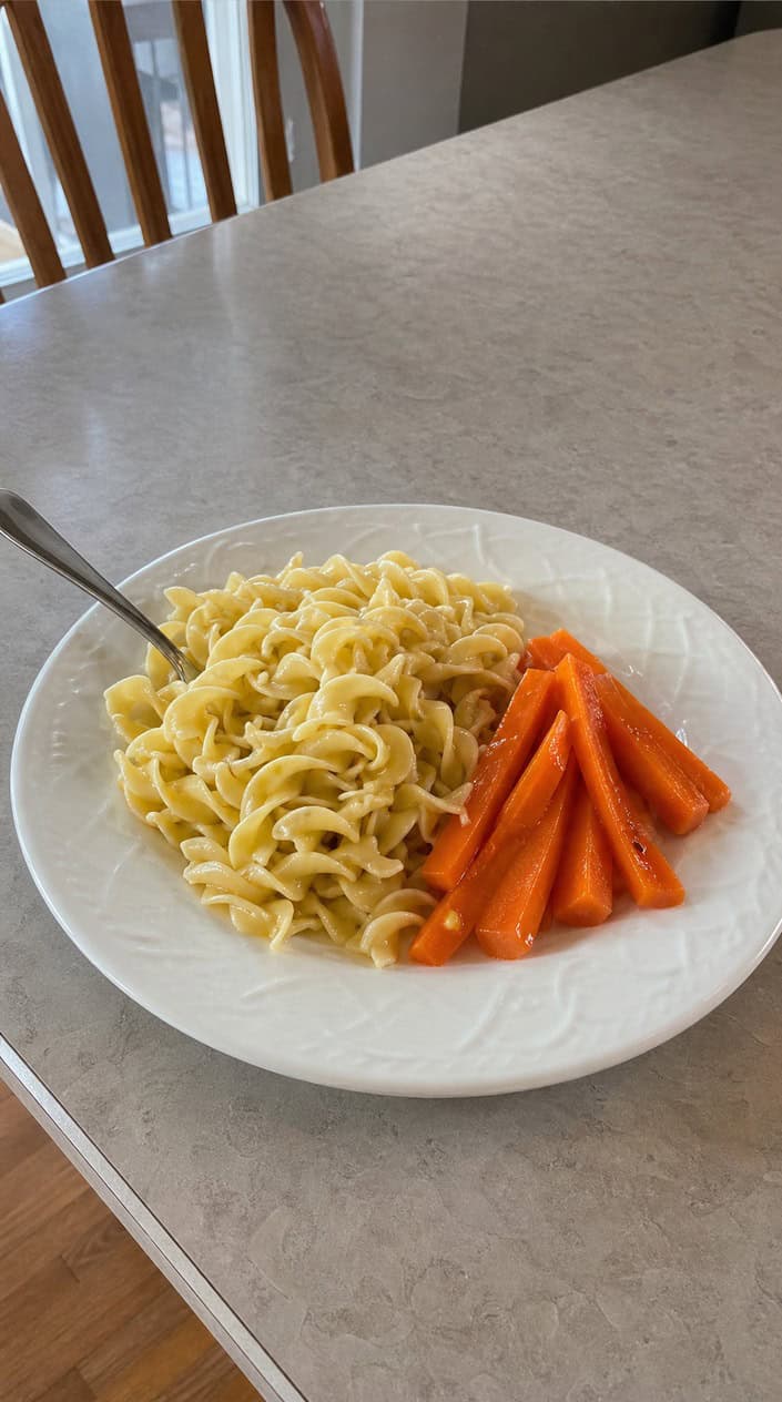 A bowl of buttered egg noodles with soft steamed orange carrot rounds arranged on top and a light honey drizzle catching the light