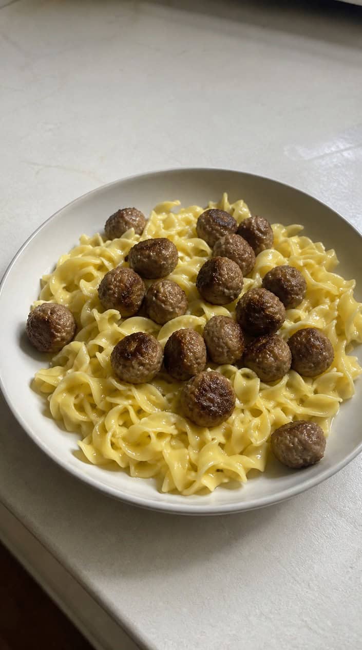 A deep bowl of buttered egg noodles topped with small browned meatballs, styled on a rustic wooden table with a linen napkin to the side