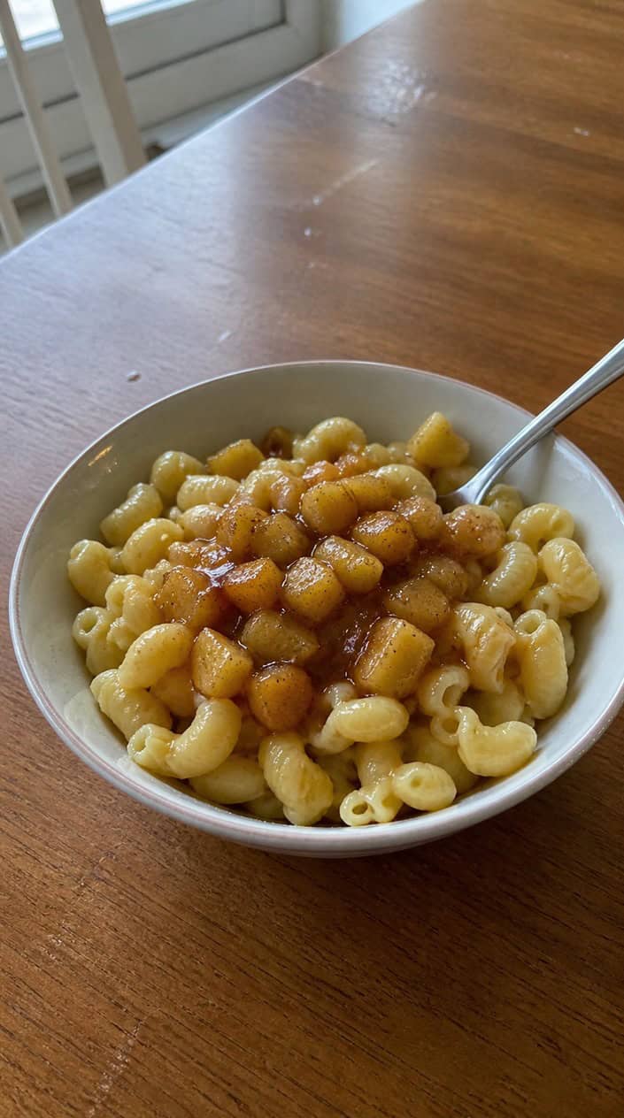 A small bowl of soft egg noodles coated in a warm amber apple-cinnamon butter sauce, with fresh apple slices and cinnamon sticks arranged nearby on a wooden surface
