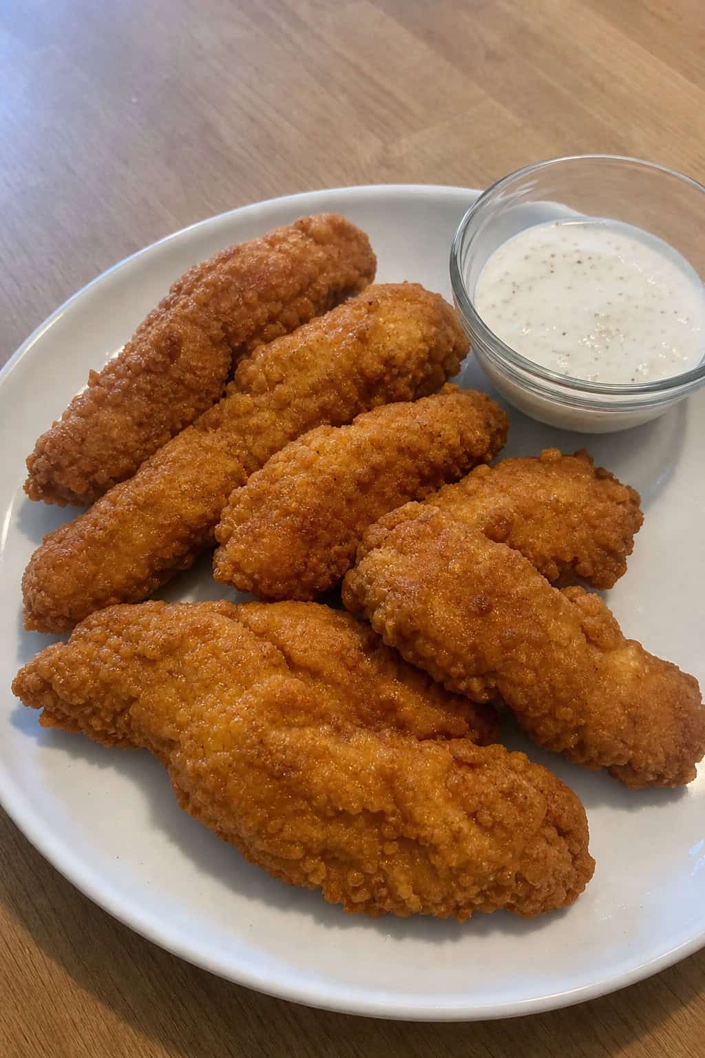 A plate of golden fried chicken tenders with fresh herbs and a bowl of dipping sauce on a wooden table.