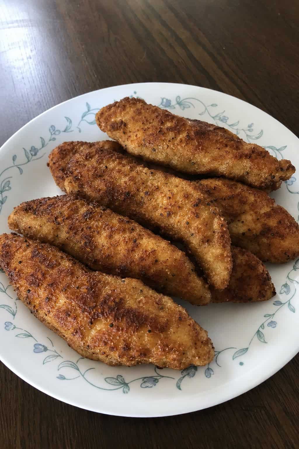 Golden-brown pan-fried chicken tenderloins on a white plate, garnished with fresh parsley and lemon wedges on a wooden table.