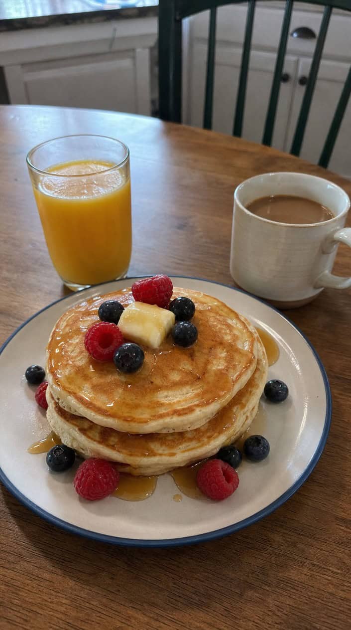 A stack of buttermilk pancakes with berries and syrup on a picnic table decorated with small American flags in a backyard setting.