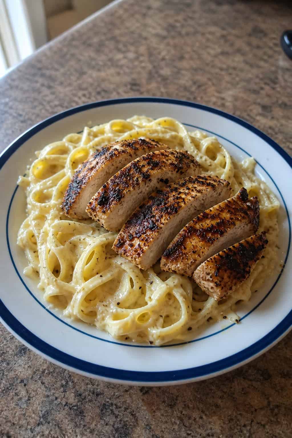 A plate of creamy Alfredo pasta topped with slices of blackened Cajun chicken, garnished with parsley and Parmesan cheese on a wooden table.