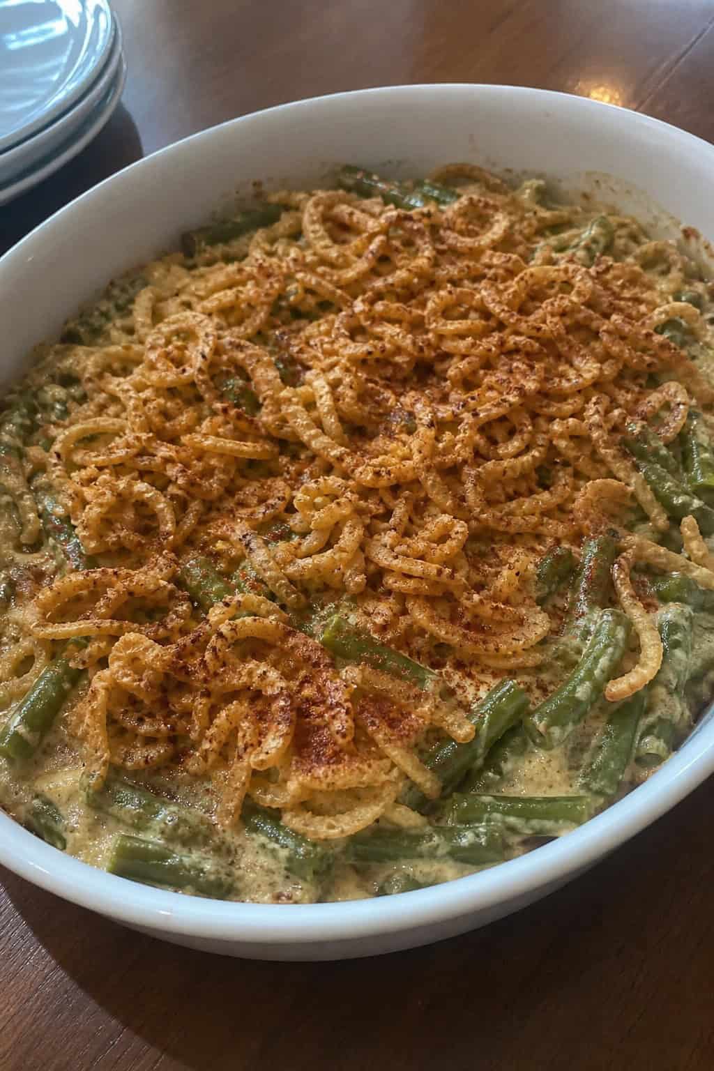A baked green bean casserole topped with golden crispy fried onions in a white ceramic dish on a wooden table, with a rustic linen napkin beside it.