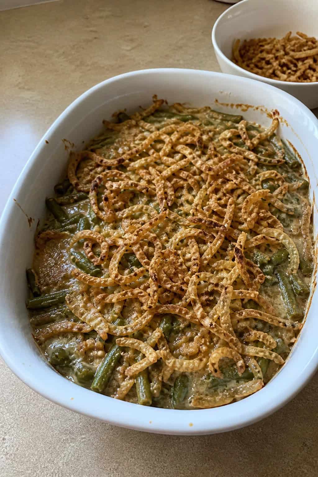 A baked green bean casserole topped with a thick, golden layer of crispy fried onions in a white ceramic dish, with fresh green beans and a wooden serving spoon beside it on a wooden table.