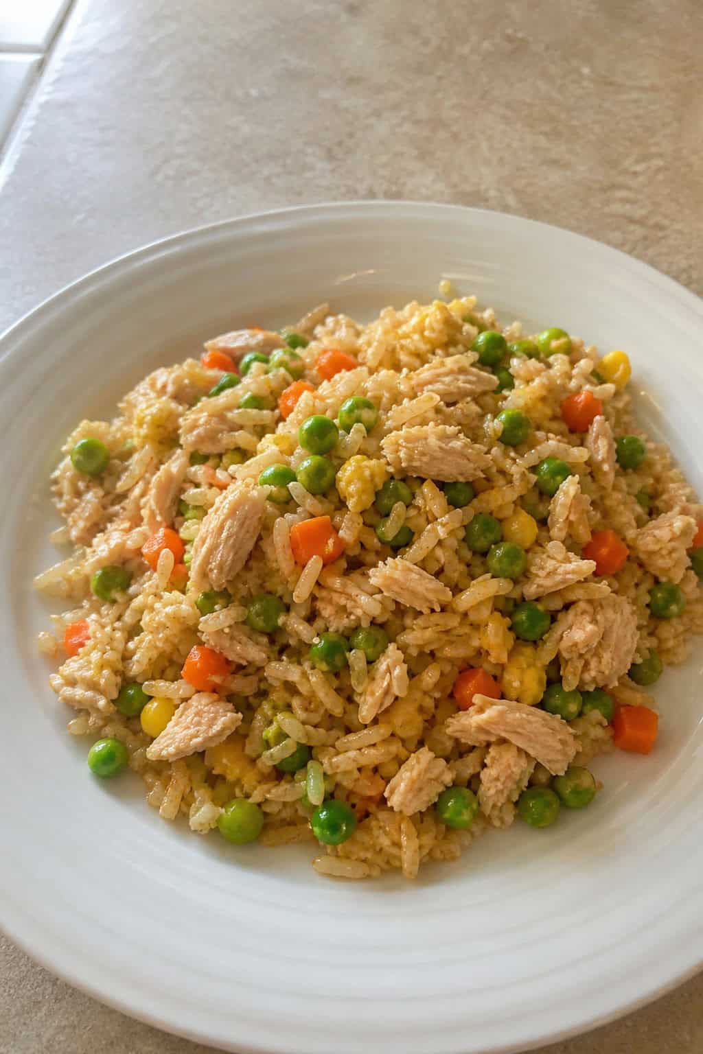 A plate of canned chicken and vegetable fried rice on a wooden table with fresh vegetables and soy sauce in the background.
