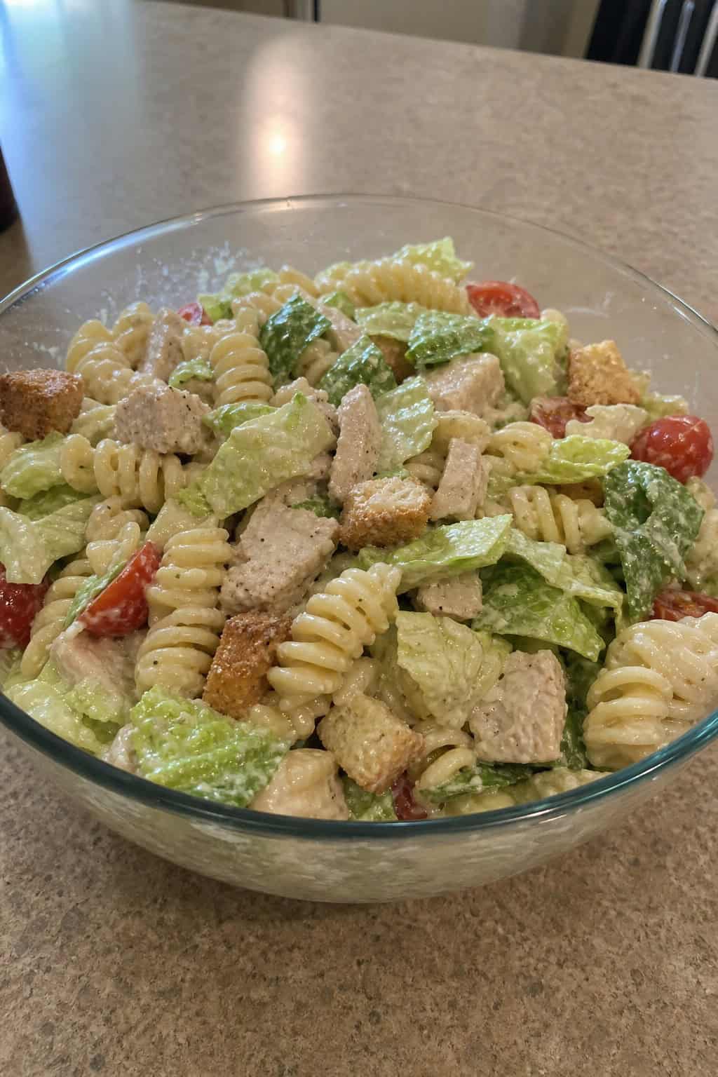 A bowl of pasta salad with canned chicken, lettuce, cherry tomatoes, Parmesan cheese, and croutons on a wooden table with a fork and small bowl of dressing nearby.