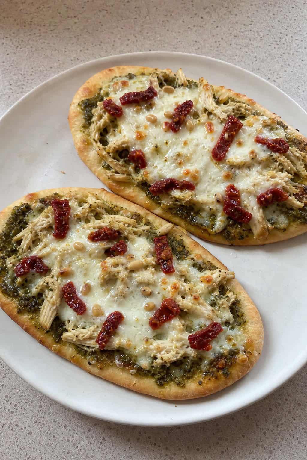 Flatbreads topped with shredded chicken, pesto, cheese, and sun-dried tomatoes on a wooden board with fresh basil and a bowl of pesto nearby.