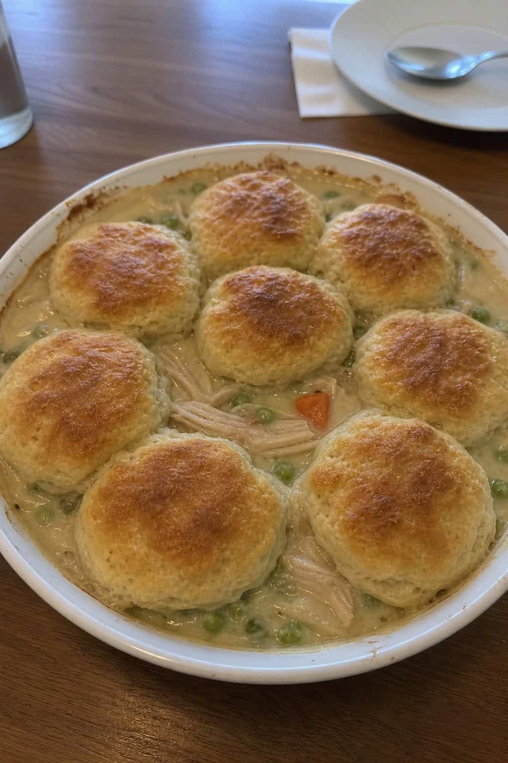 A freshly baked chicken pot pie topped with golden biscuits in a ceramic dish on a wooden table, surrounded by fresh vegetables and herbs.