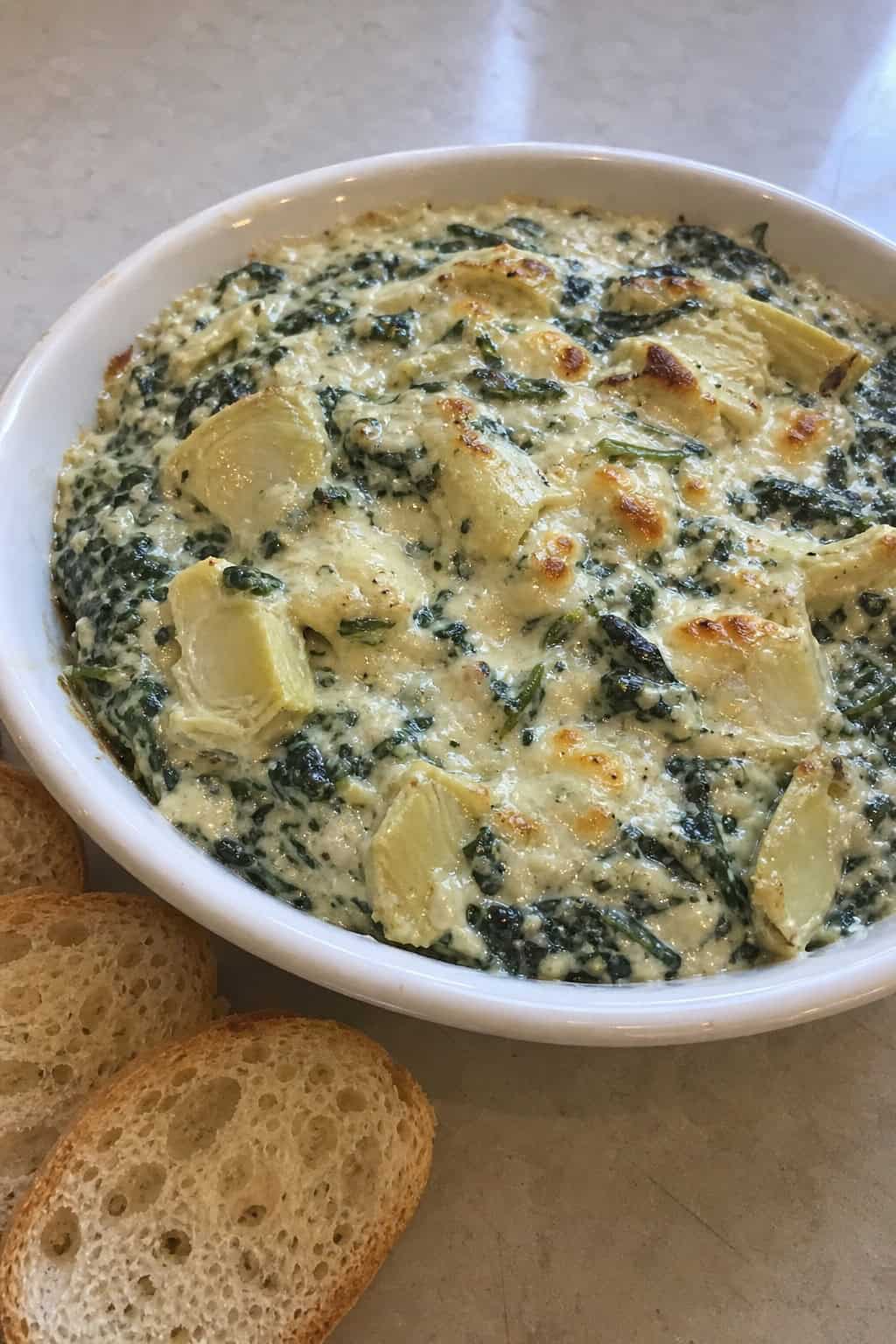 A bowl of creamy spinach artichoke dip with chunks of chicken, surrounded by sliced bread and vegetable sticks on a wooden board.