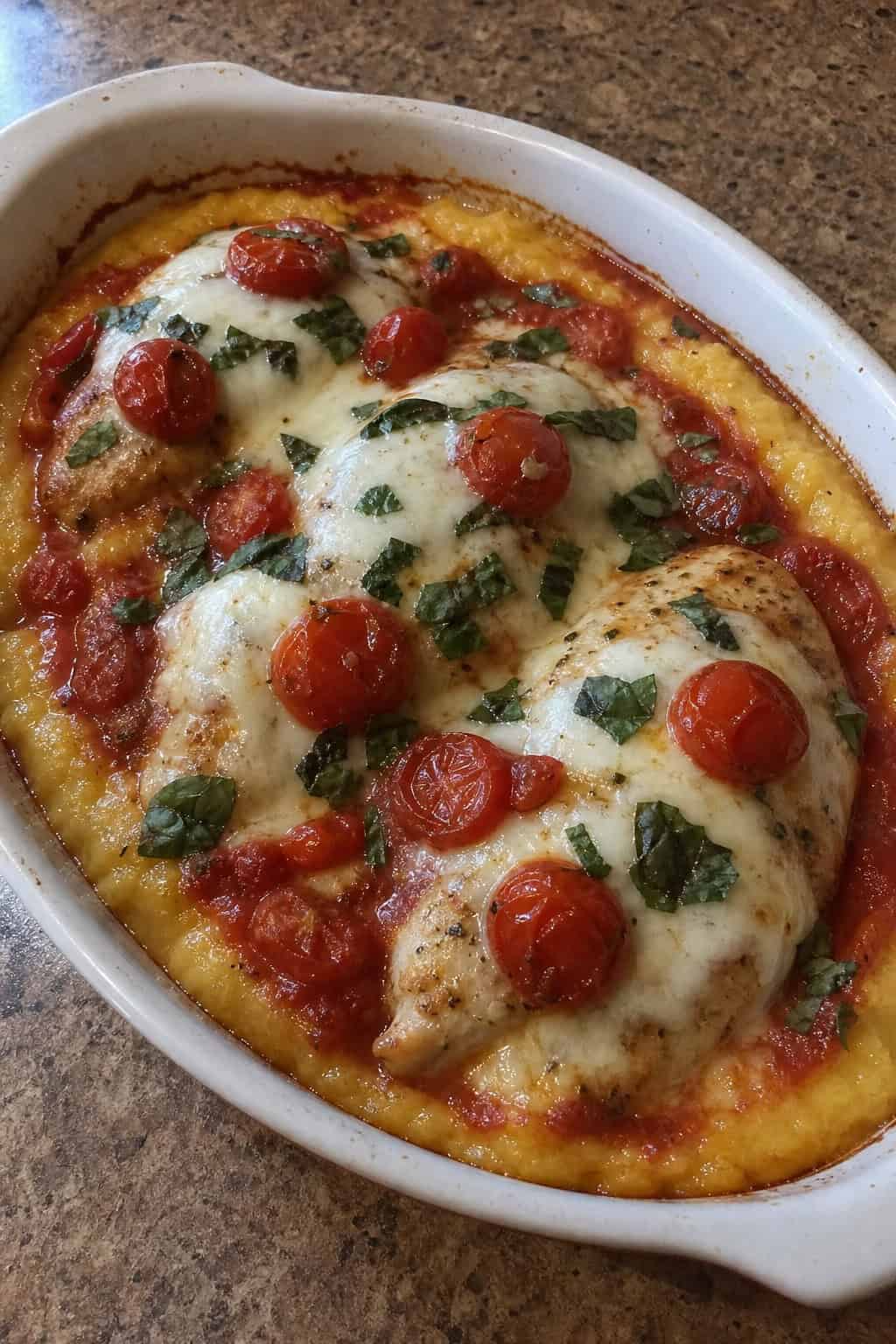 A baked Caprese chicken and polenta casserole in a ceramic dish with melted mozzarella, blistered cherry tomatoes, and fresh basil leaves on a wooden table.