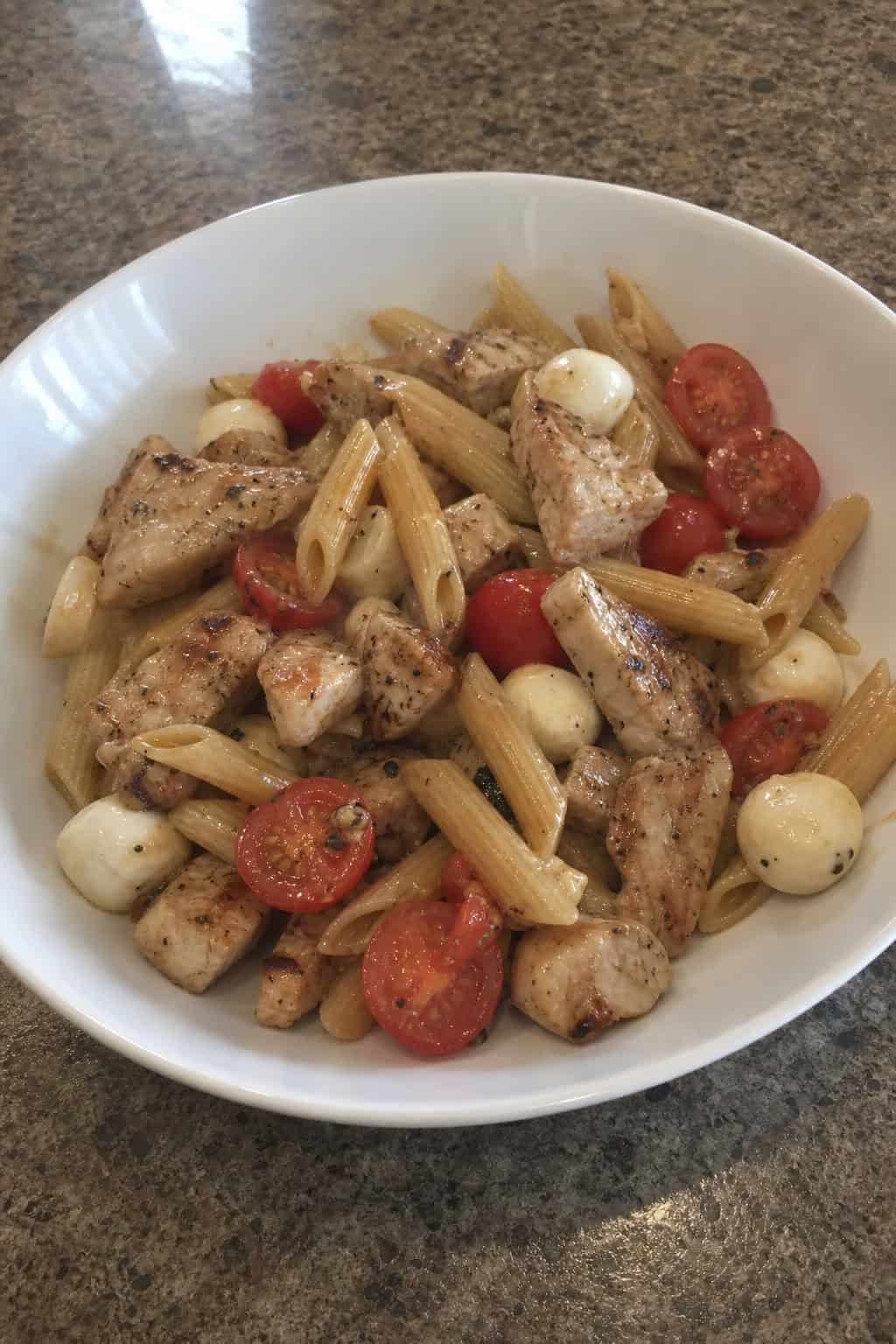 A bowl of Caprese chicken pasta with chicken pieces, pasta, cherry tomatoes, basil, and mozzarella on a wooden table.