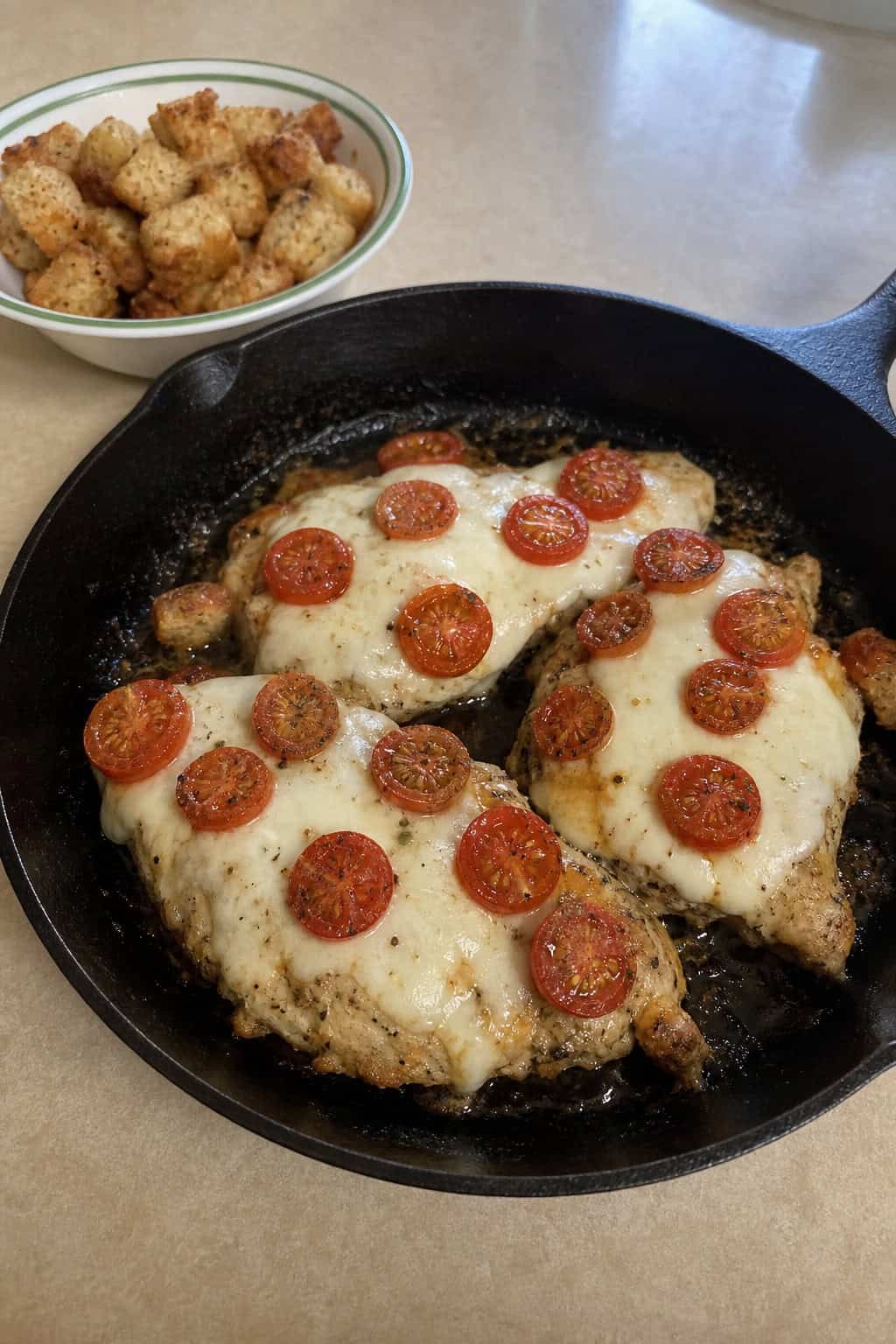 A skillet with Caprese chicken topped with melted cheese, basil, and cherry tomatoes, surrounded by homemade croutons on a wooden table.