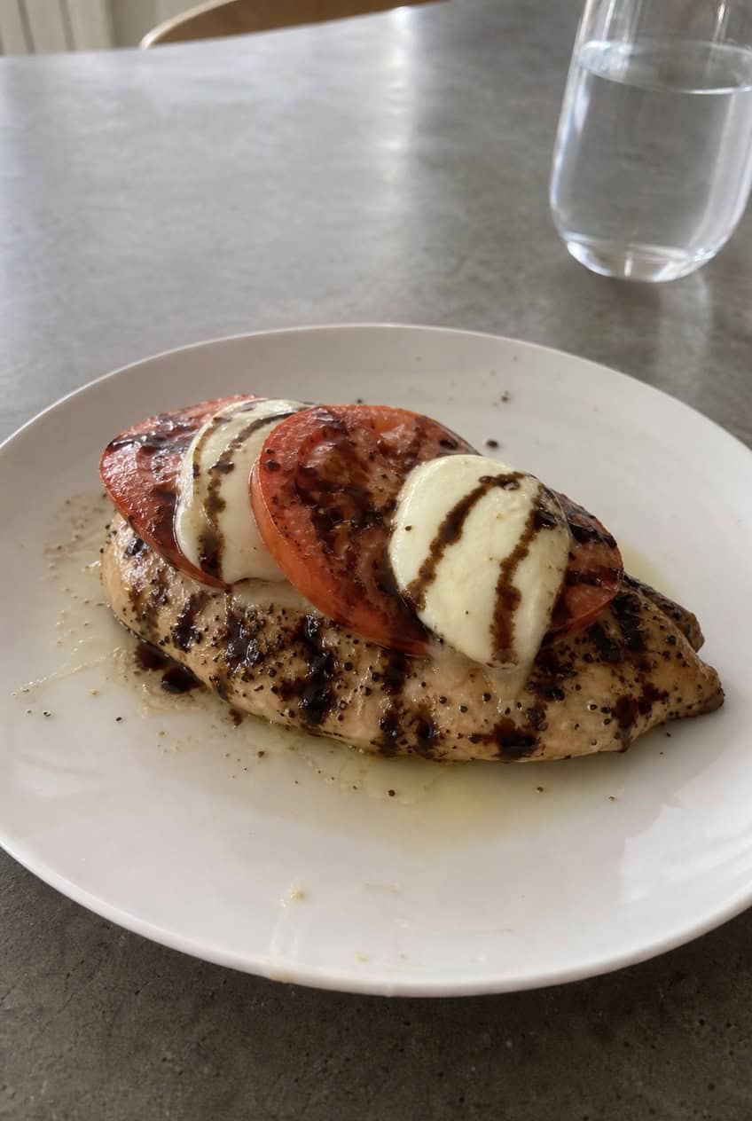 A white baking dish with shredded rotisserie chicken layered under rounds of fresh mozzarella and ripe tomato slices, finished with a dark balsamic drizzle and torn fresh basil leaves, on a wooden table.