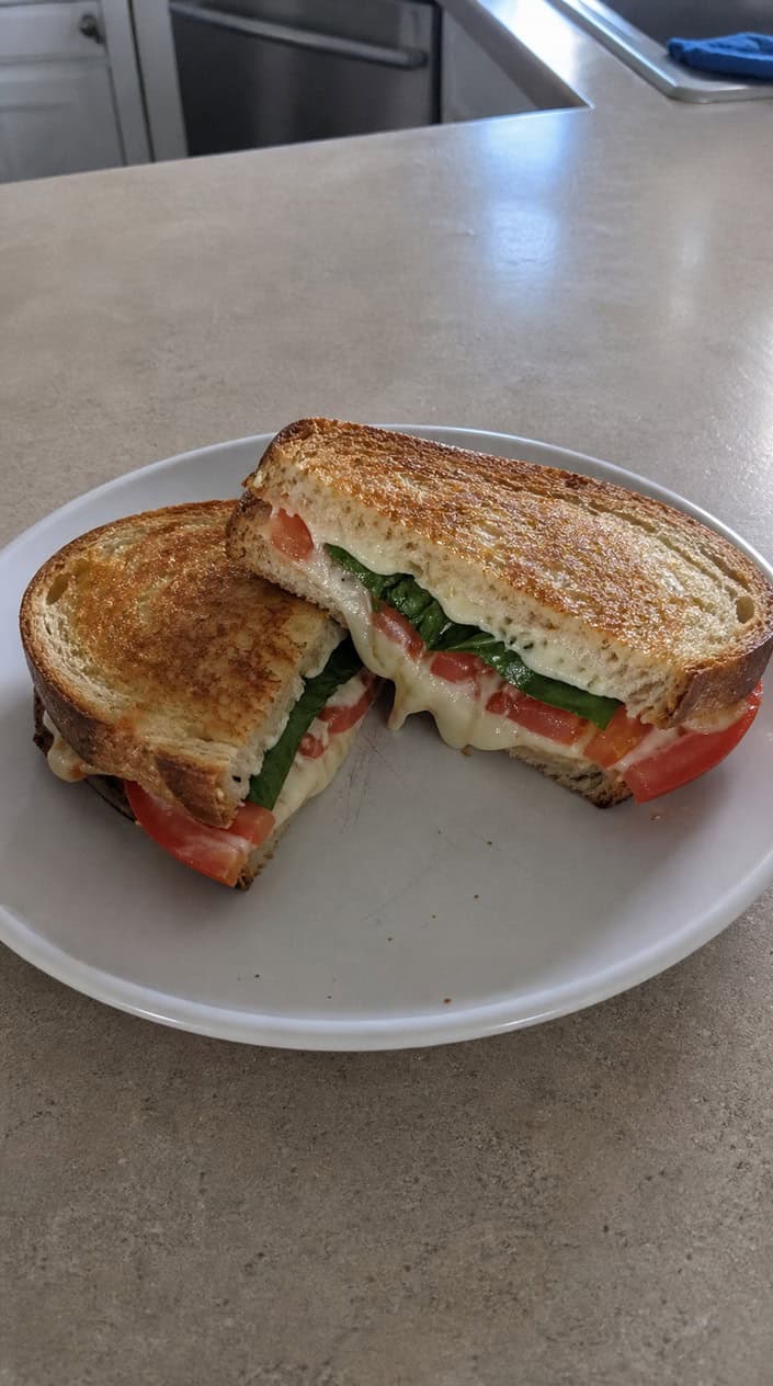 A Caprese grilled cheese sandwich on sourdough cut in half to show the pull of melted mozzarella, a slice of ripe tomato, and bright green basil, resting on a wooden board with a small drizzle of balsamic nearby.