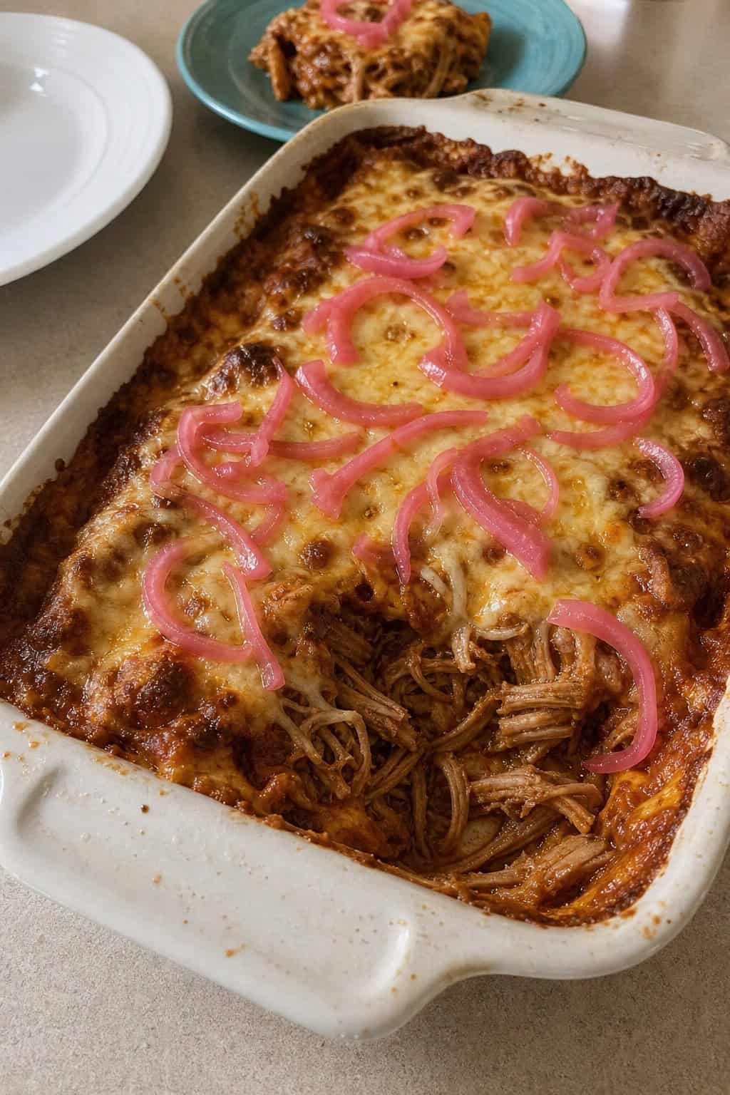 A baked carnitas enchilada casserole in a ceramic dish topped with bright pink pickled red onions and fresh cilantro, with small ramekins of salsa and lime wedges arranged on the wooden table beside it.