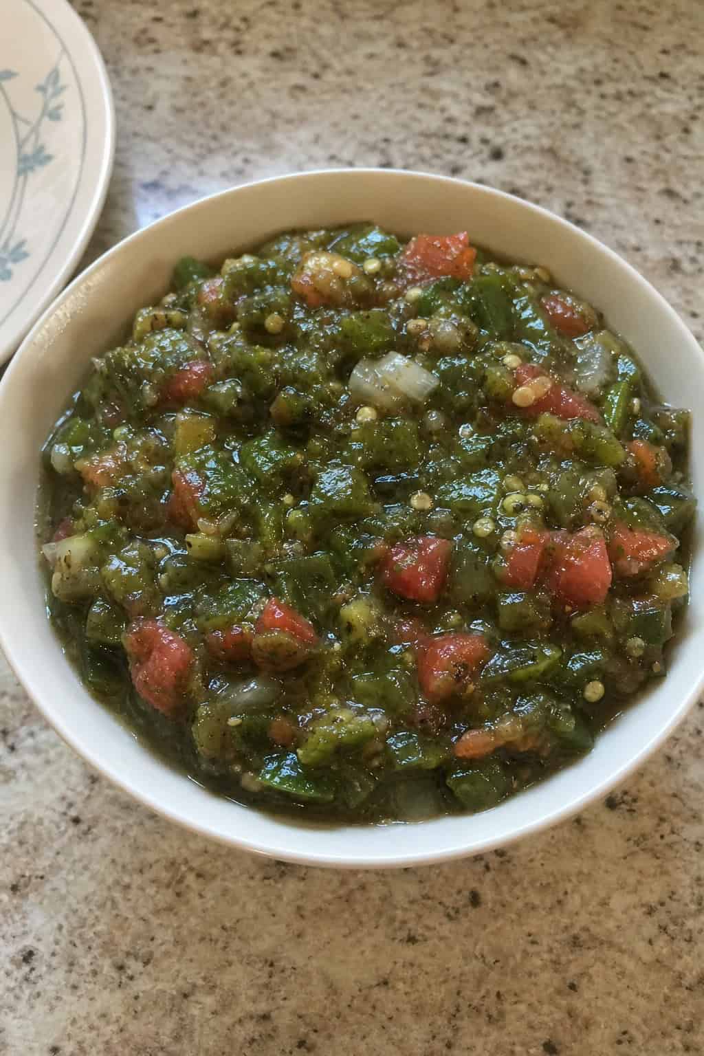 A bowl of charred poblano pepper salsa on a wooden table surrounded by fresh poblano peppers, lime wedges, garlic, and sea salt.