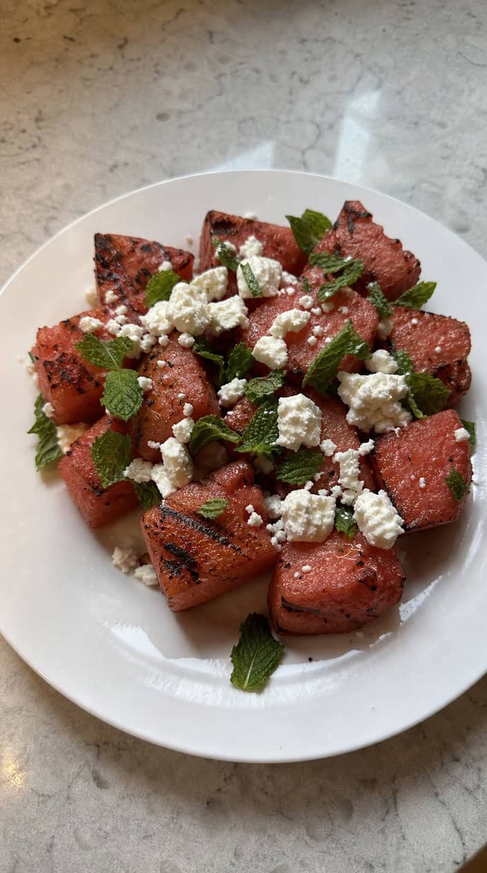 A bowl of charred watermelon, feta, and mint salad on a wooden table with summer decorations in the background.