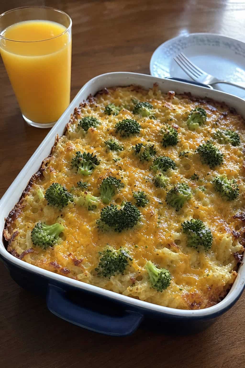 A baked cheddar broccoli hashbrown casserole in a white ceramic dish with soft broccoli florets and melted cheese visible throughout, on a wood table with a small head of fresh broccoli and a block of cheddar nearby.
