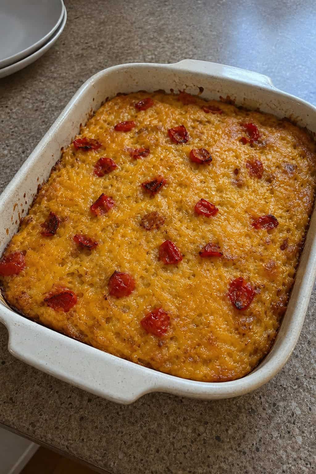 A baked cheddar and roasted red pepper hashbrown casserole in a ceramic dish on a wooden table with kitchen utensils nearby.