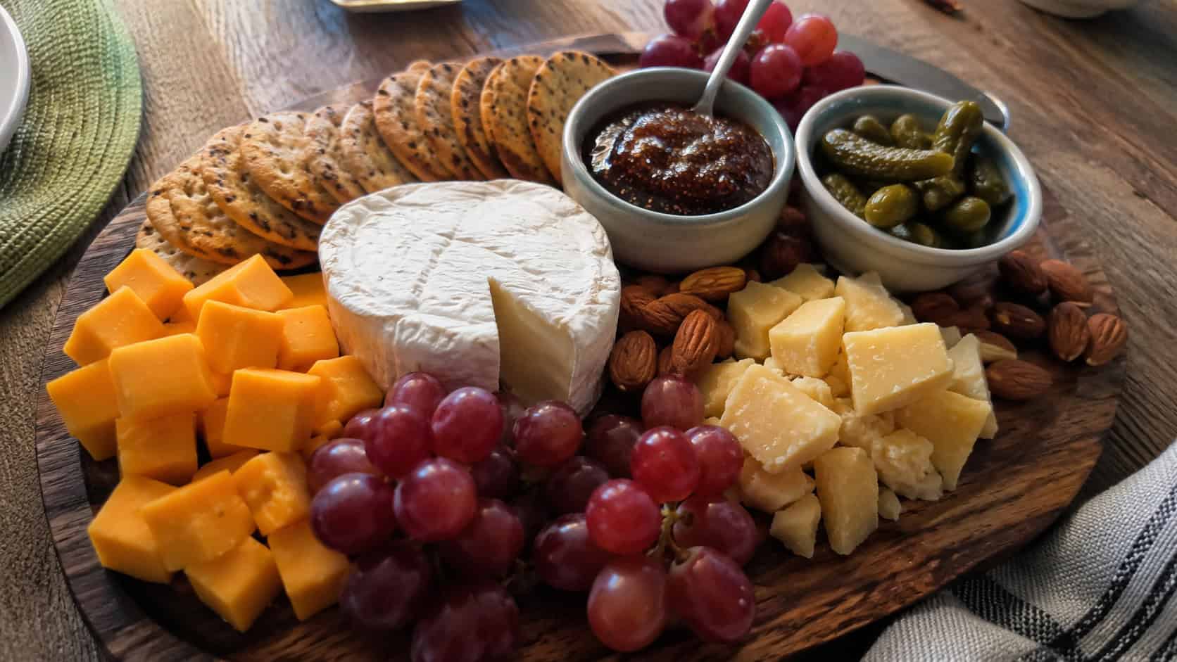 a finished cheese and cracker board on a wooden surface, cubed sharp cheddar, a wheel of brie, shards of aged gouda, fanned crackers, red grape clusters, almonds, a small bowl of fig jam and cornichons, warm natural light, casual home entertaining