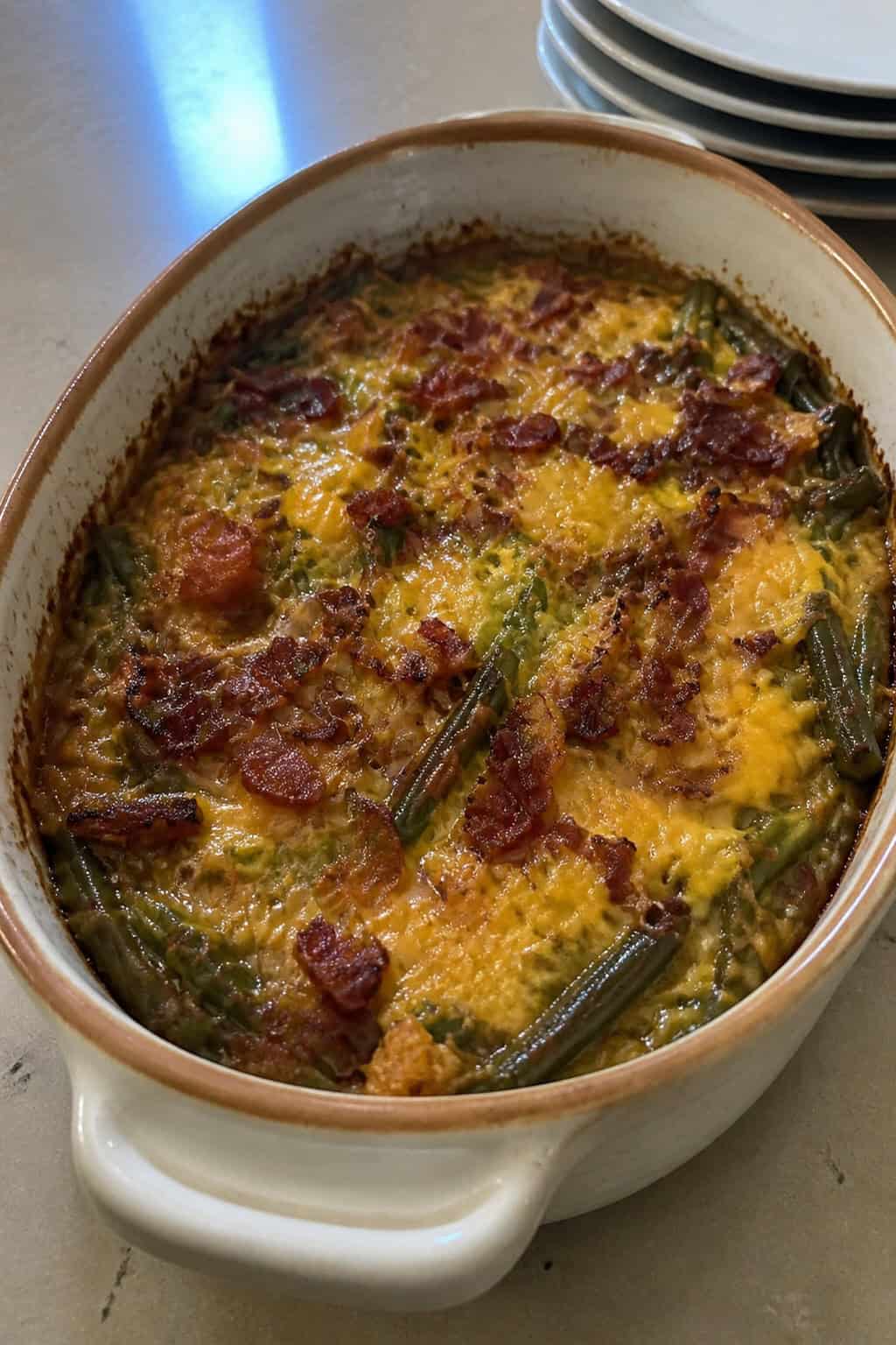 A golden baked green bean casserole with bacon and melted cheese visible through the crispy topping, in a white ceramic dish with a serving spoon resting beside it on a wooden table.